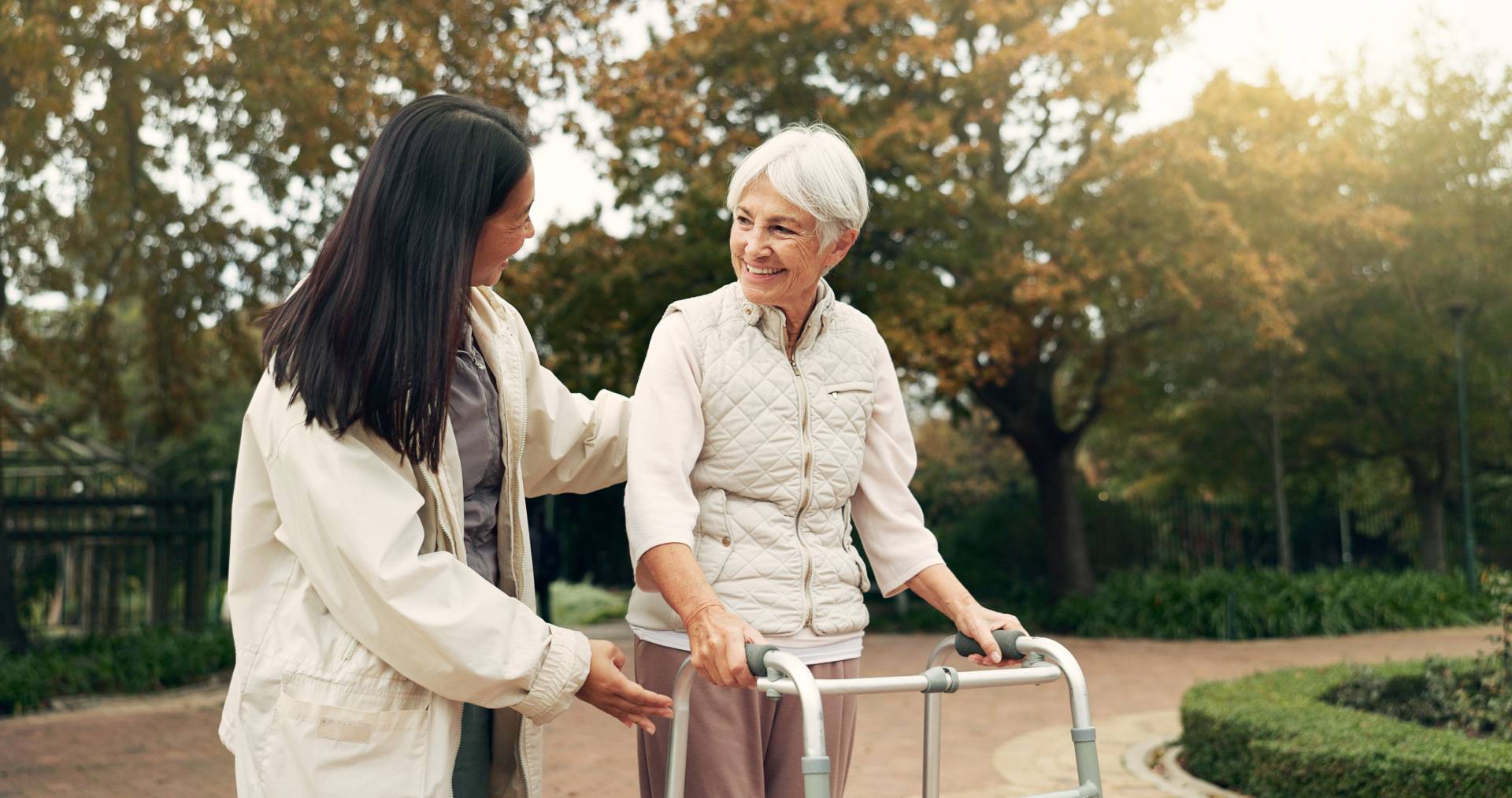 Caregiver supporting an elderly woman using a walker while walking outdoors in a park, both smiling and enjoying the moment.