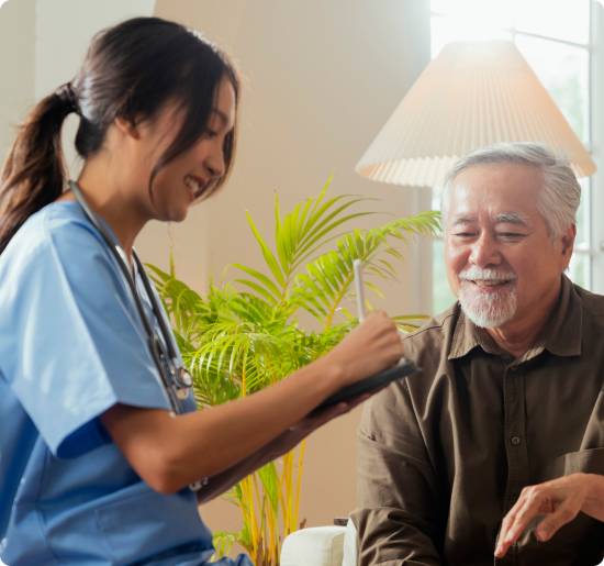 Smiling caregiver in blue scrubs taking notes on a tablet while chatting with a senior man at home