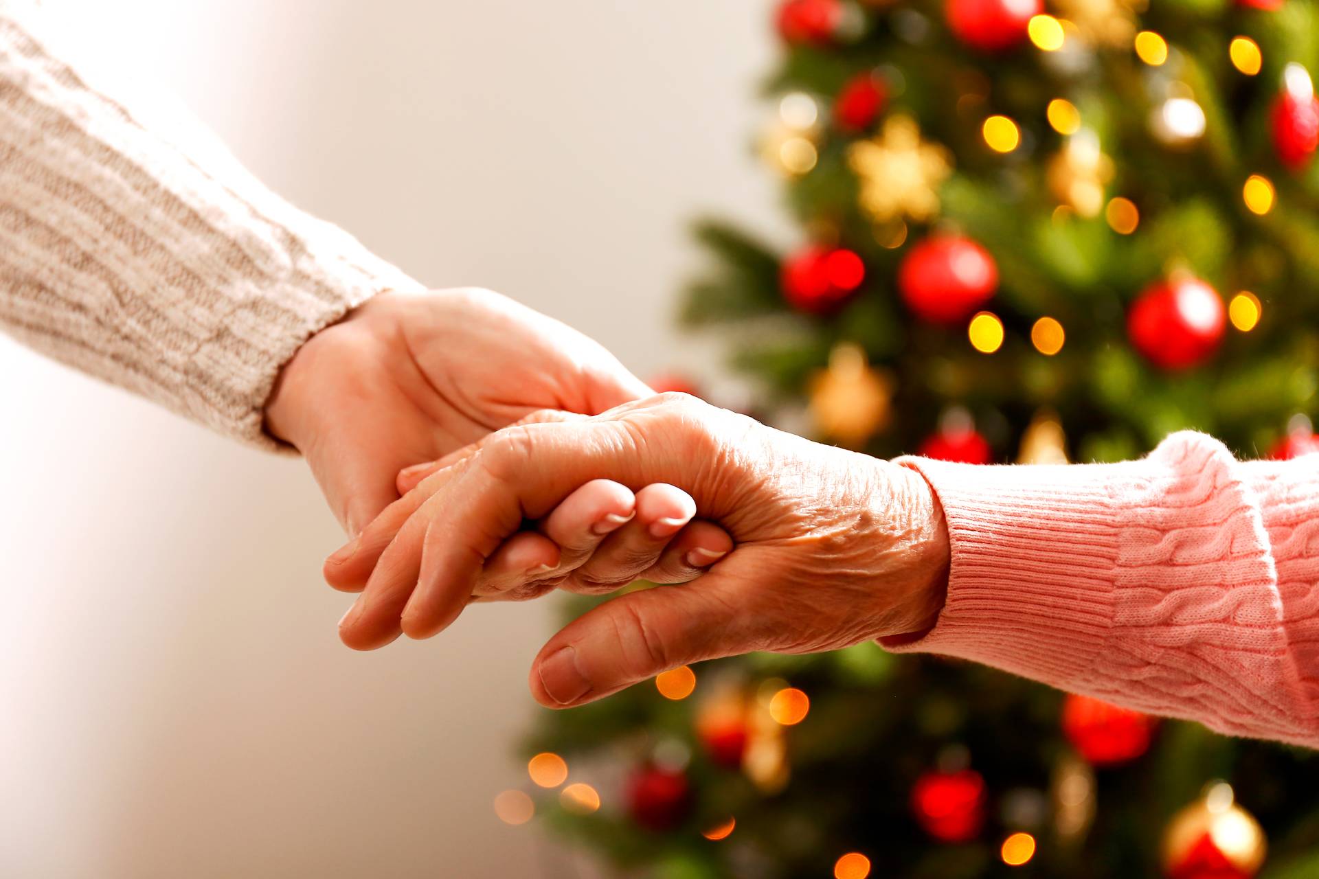 A young person holding an elderly persons hand with a Christmas tree sparkling in the background.