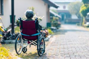 An elderly woman in a wheelchair, wearing a patterned shirt, looks at the pathway ahead. She is outdoors, with a clear view of a quiet, sunlit street lined with greenery and homes in the background.