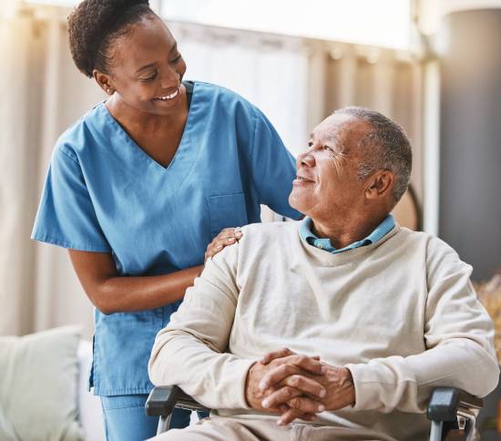 Dedicated Caregivers for Every Overnight Hour-image A smiling caregiver in blue scrubs gently rests her hand on the shoulder of an elderly man in a wheelchair. They share a warm, caring moment.