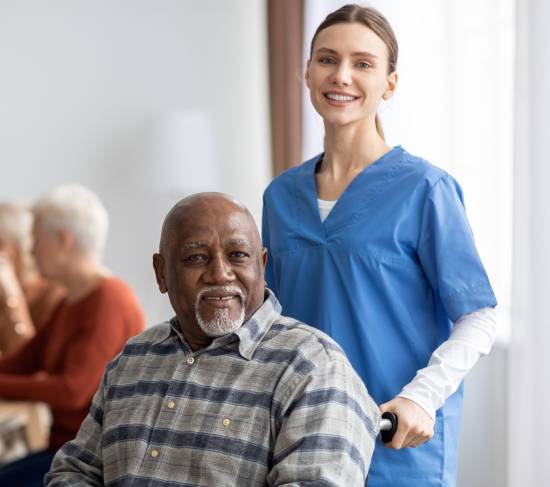 A smiling nurse in blue scrubs stands beside an elderly man in a wheelchair, conveying warmth and care. Other seniors are visible in the background.