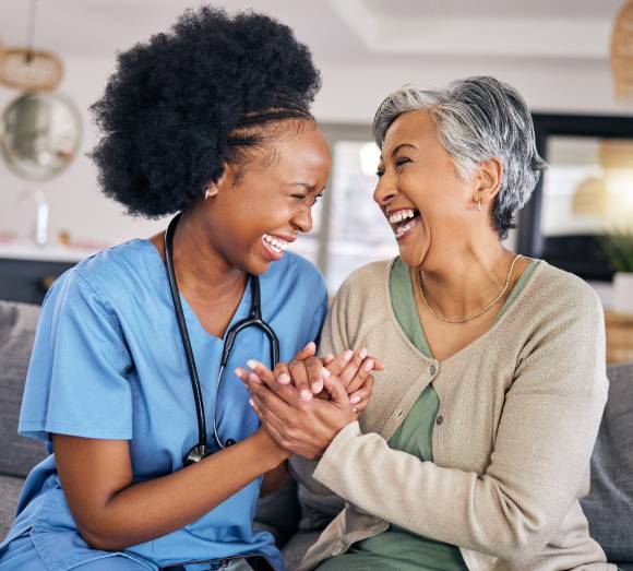 Providing Peace of Mind for Edmonton Families-image A nurse in blue scrubs shares a heartfelt laugh with an elderly woman on a couch. They hold hands warmly, conveying joy and connection in a light-filled room.