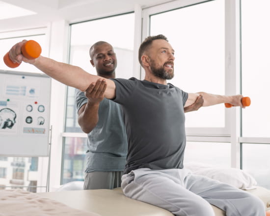 What_is_Traumatic_Brain_Injury_Home_Care__image-0bKK4rjs_optimzied A man sits on a therapy table, arms extended with orange dumbbells, assisted by a therapist. They are in a bright room with large windows, conveying focus and support.
