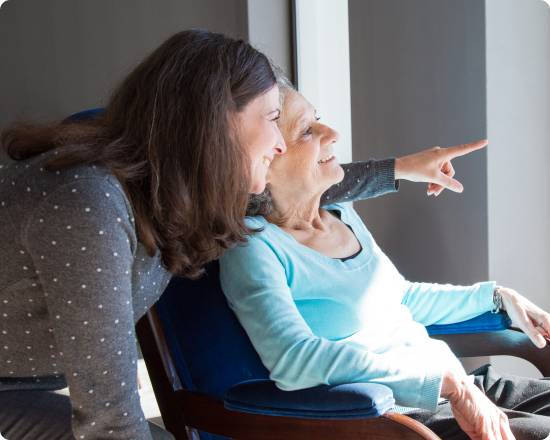 Caregiver and senior woman sitting by a window, smiling and pointing outside together