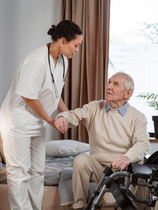Your_Trusted_Provider_for_Edmonton_TBI_Care_at_Home_image-VdYV4rjs_optimized A caregiver assists an elderly man in a cozy room. The man sits on a bed, holding a walker. The caregiver is supportive, creating a warm, caring atmosphere.