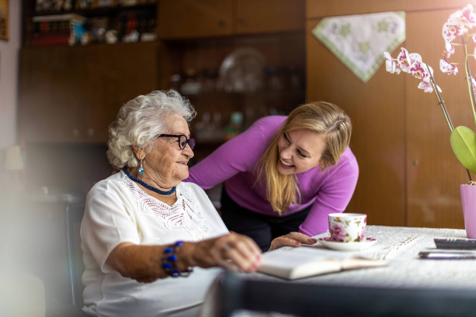 Smiling caregiver talking with an elderly woman at a table, sharing a warm moment together with a book and teacup in front of them.