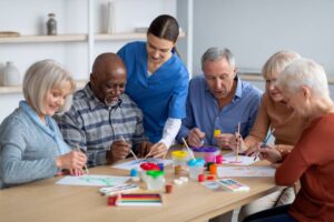 Caregiver helps a group of older adults paint together at a table with art supplies.