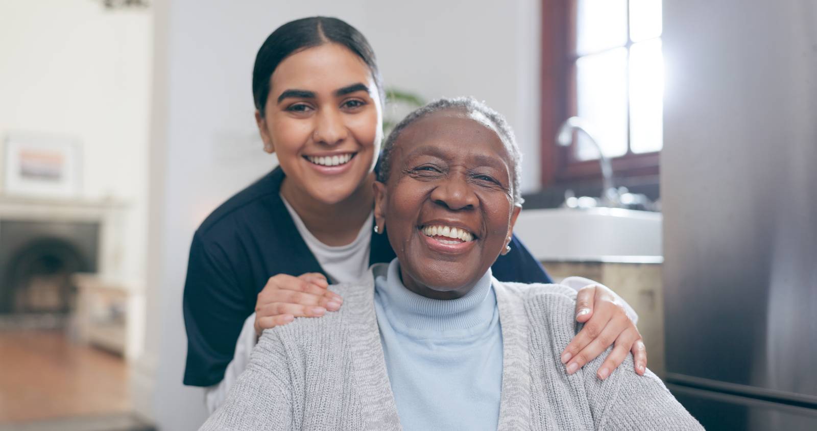 A joyful woman seated while a caregiver stands behind her with her hands on the elder's shoulders.