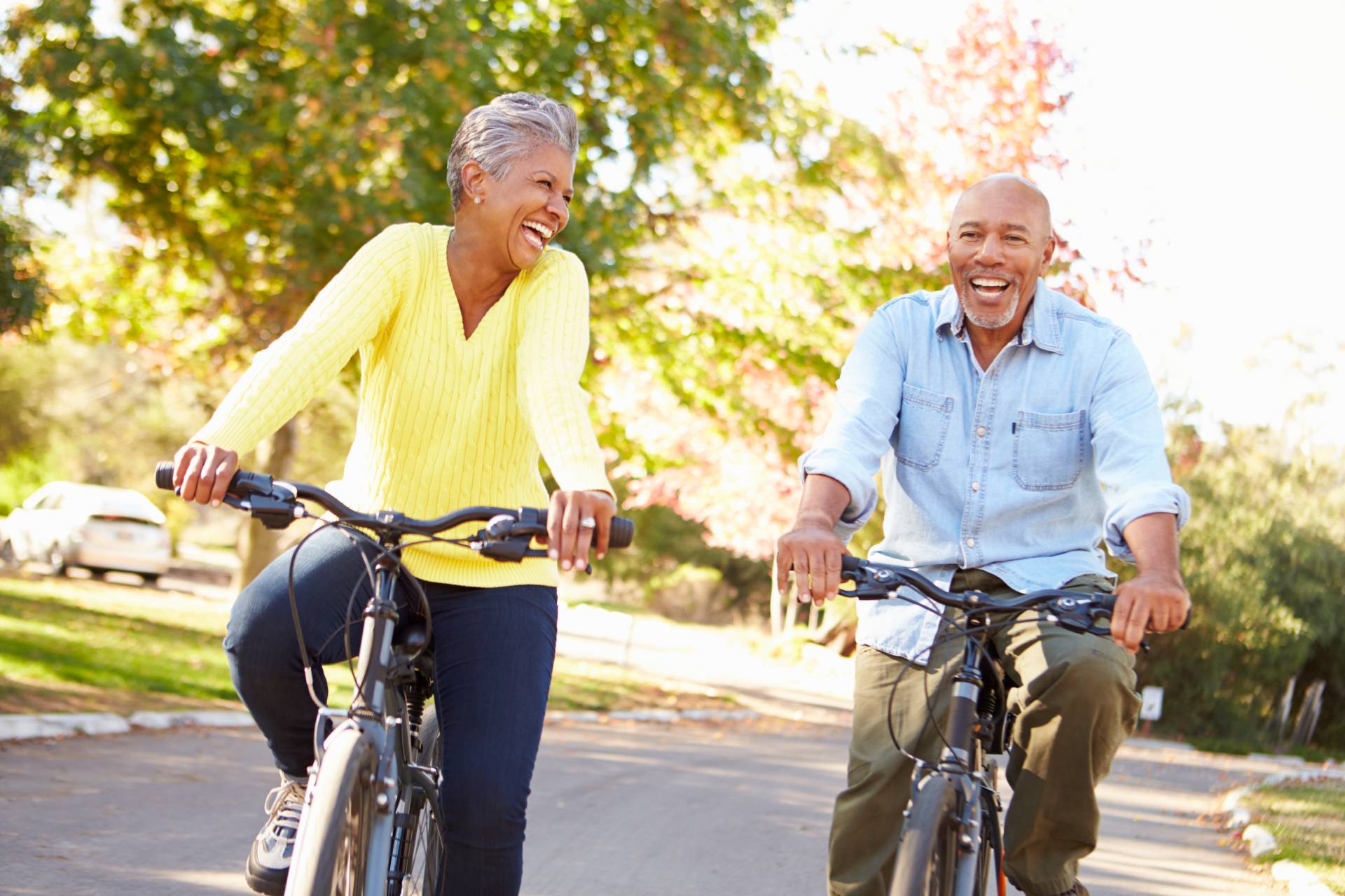 Two older adults laughing while riding bicycles on a sunny neighborhood street.
