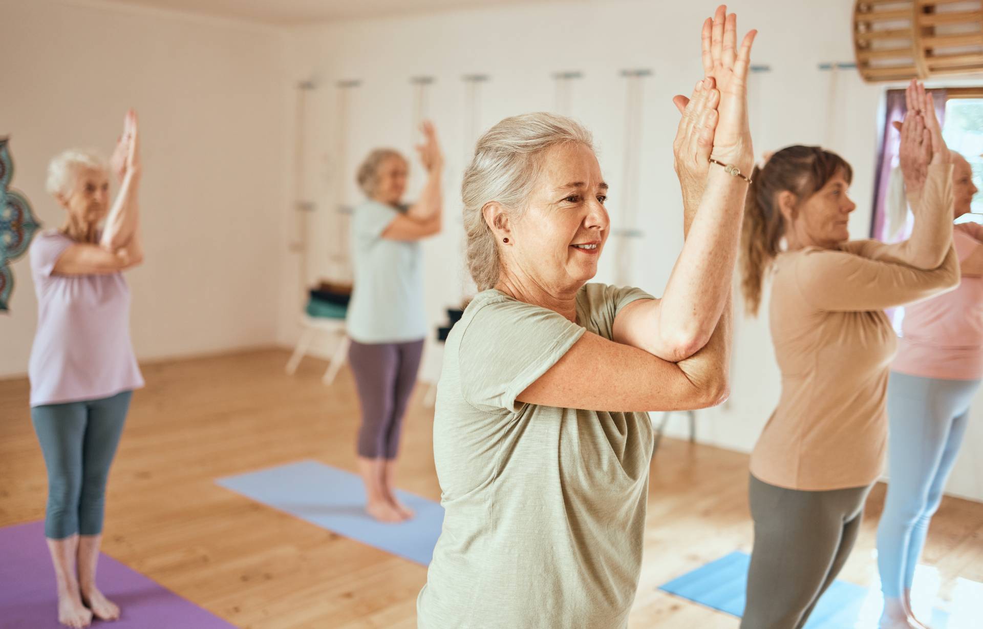 Older adults doing a gentle yoga class on mats in a studio, raising their arms in a stretch.