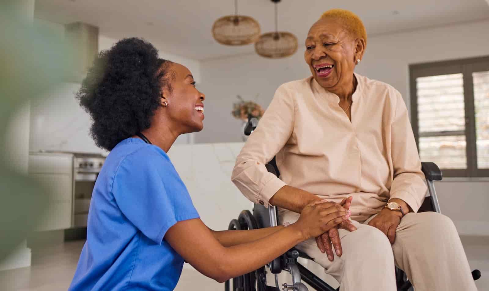 smiling caregiver in blue scrubs kneeling beside an older woman in a wheelchair, holding her hands and sharing a joyful conversation in a bright, modern home setting.