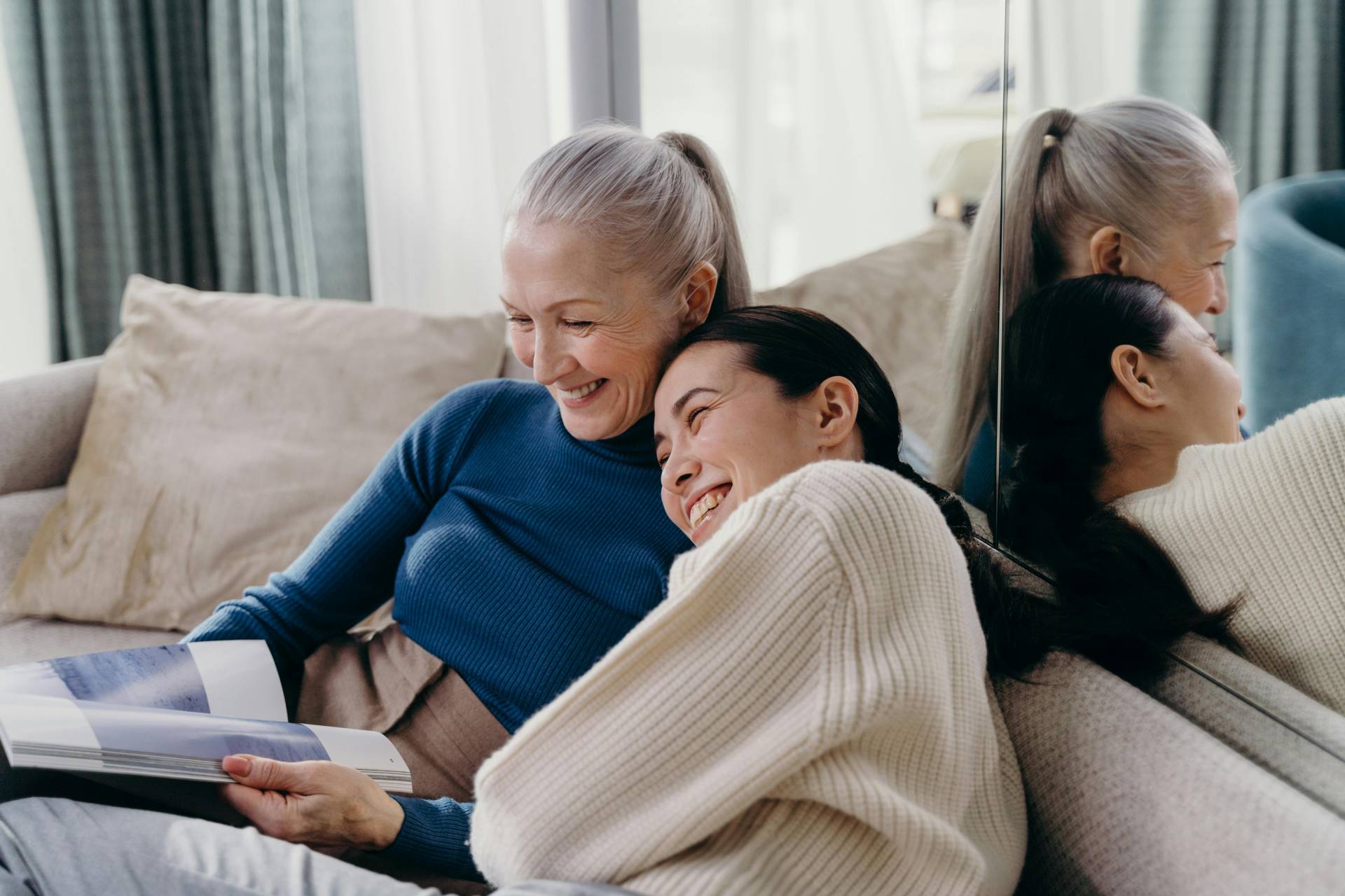 A smiling elderly woman and a younger woman are sitting on a couch, sharing a moment while looking at a photo album together.