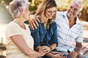 An elderly couple is sitting on a bench outdoors with their adult daughter. The elderly man has his arm around the daughter, and all three are smiling and laughing together, sharing a joyful moment in the sunlight.