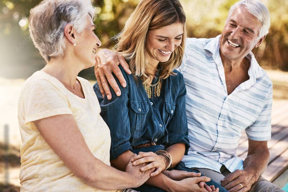 An elderly couple is sitting on a bench outdoors with their adult daughter. The elderly man has his arm around the daughter, and all three are smiling and laughing together, sharing a joyful moment in the sunlight.