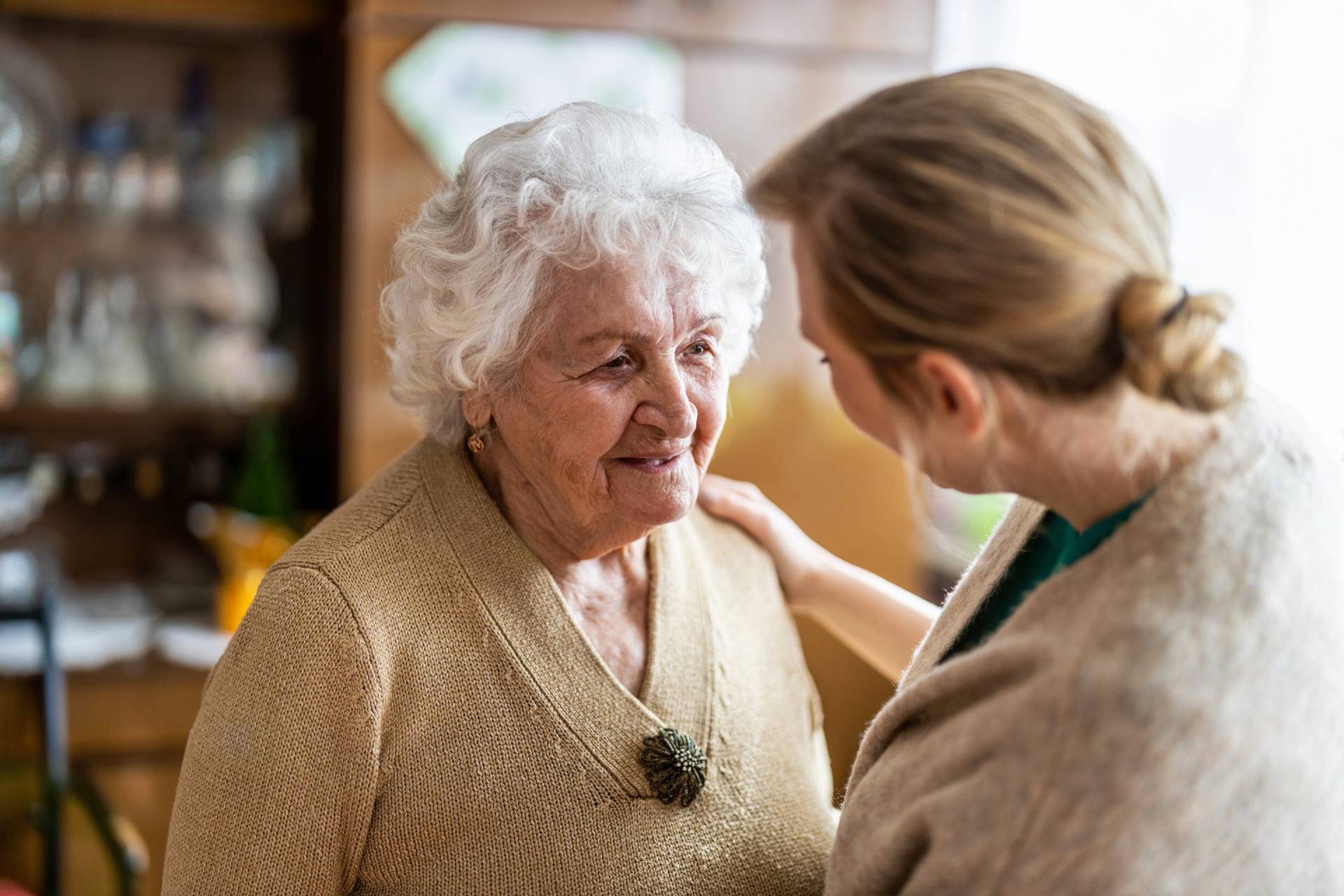 Elderly woman with a soft smile, wearing a beige sweater, engaging warmly with a caregiver who is offering supportive conversation.