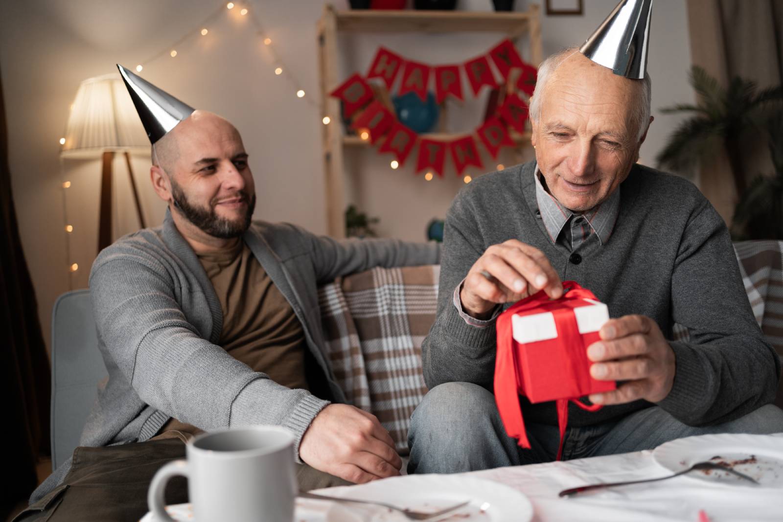 An elderly man wearing a party hat and gray sweater, opening a small gift wrapped with a red ribbon, while a younger man in a gray cardigan sits beside him, smiling.