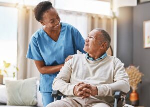 Smiling home care nurse in blue scrubs standing beside an elderly man in a wheelchair, gently resting her hand on his shoulder as they look at each other in a bright living room.
