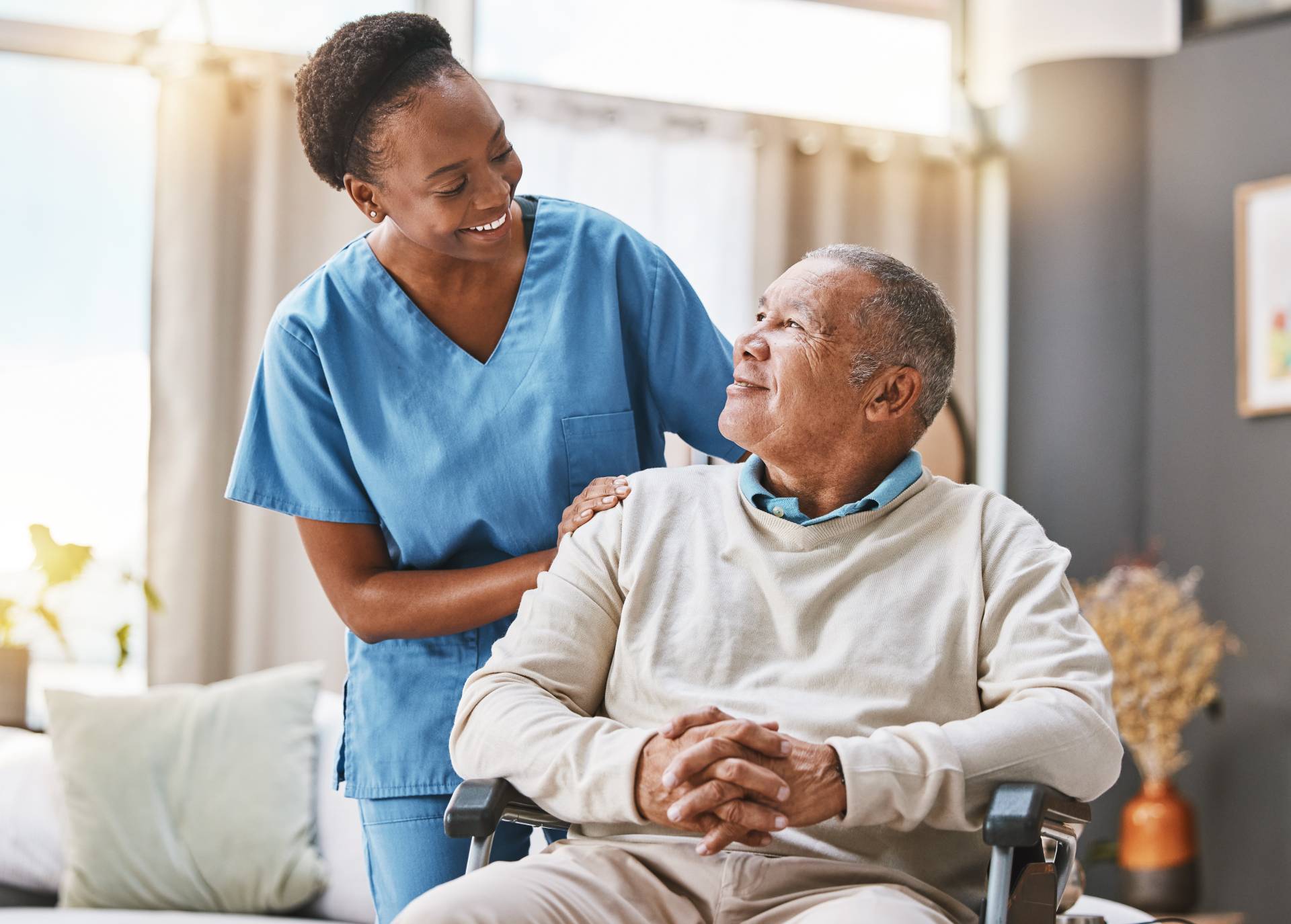 Smiling home care nurse in blue scrubs standing beside an elderly man in a wheelchair, gently resting her hand on his shoulder as they look at each other in a bright living room.