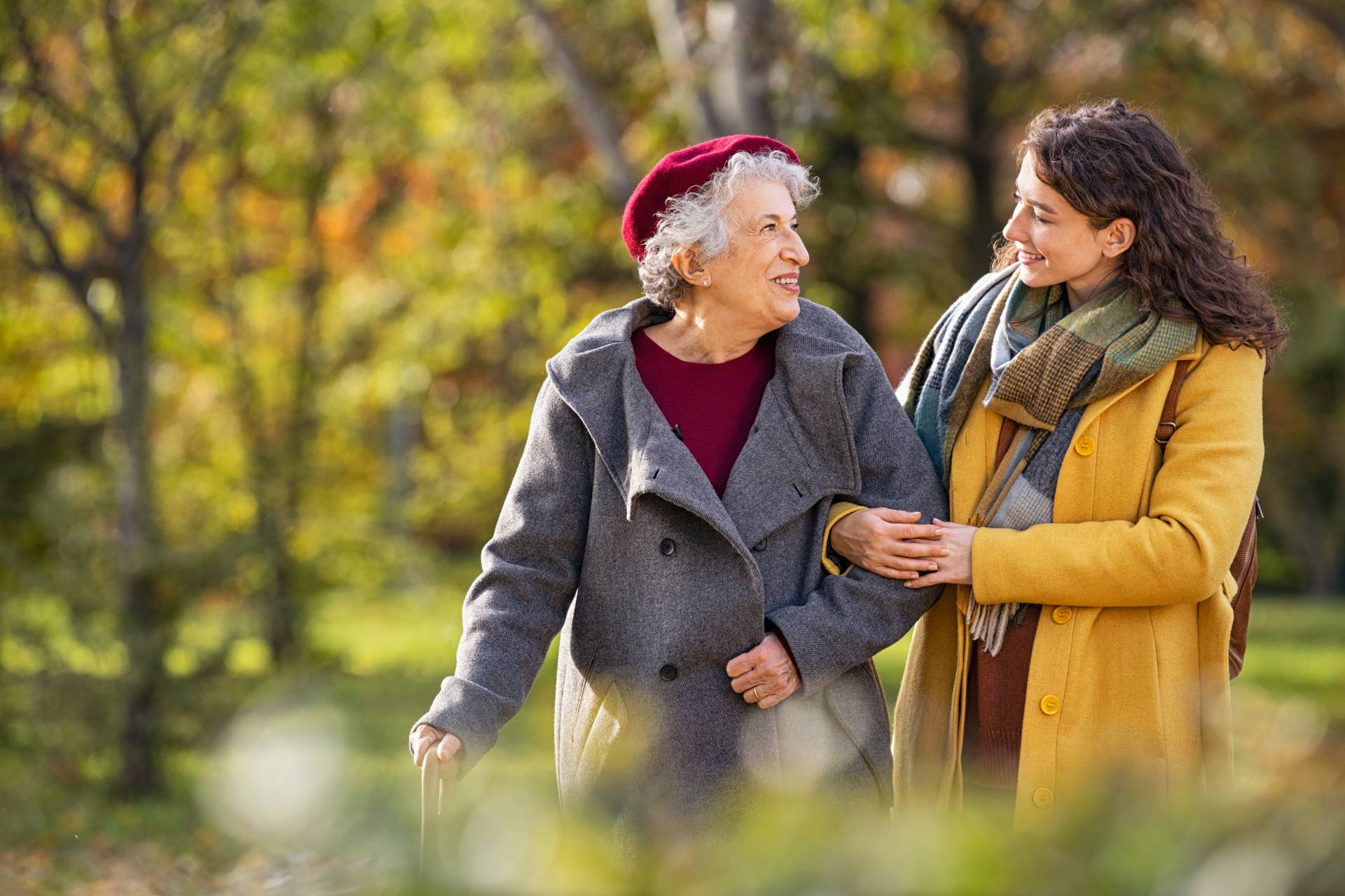 An elderly woman walking arm-in-arm with a younger woman, both smiling at each other while strolling outdoors on an autumn day.