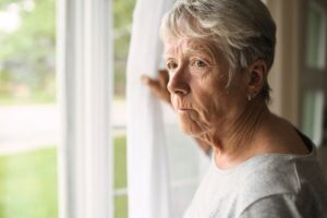 An elderly woman with short gray hair wearing a grey shirt gazing thoughtfully out of a window.