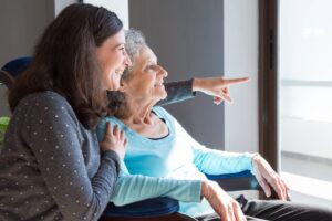 A smiling elderly woman in a wheelchair points out a window, sharing a joyful moment with a younger woman beside her.