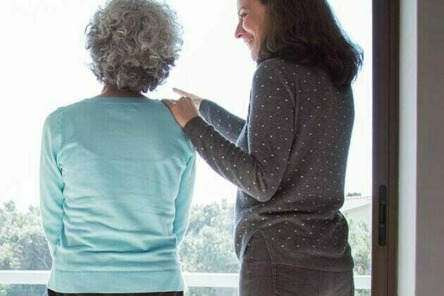 Elderly woman and her caregiver standing and looking out of a window.