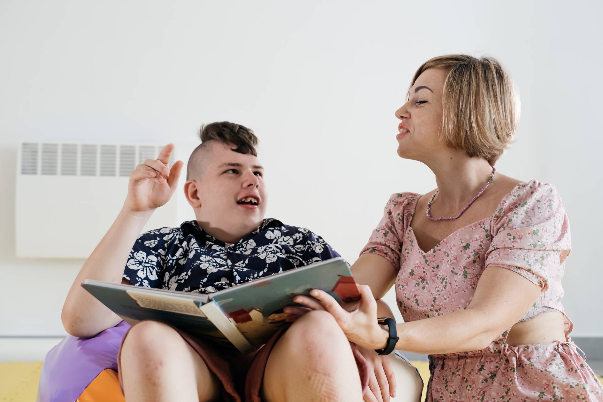 A caregiver reads a book to a child with special needs who is sitting comfortably in a beanbag.