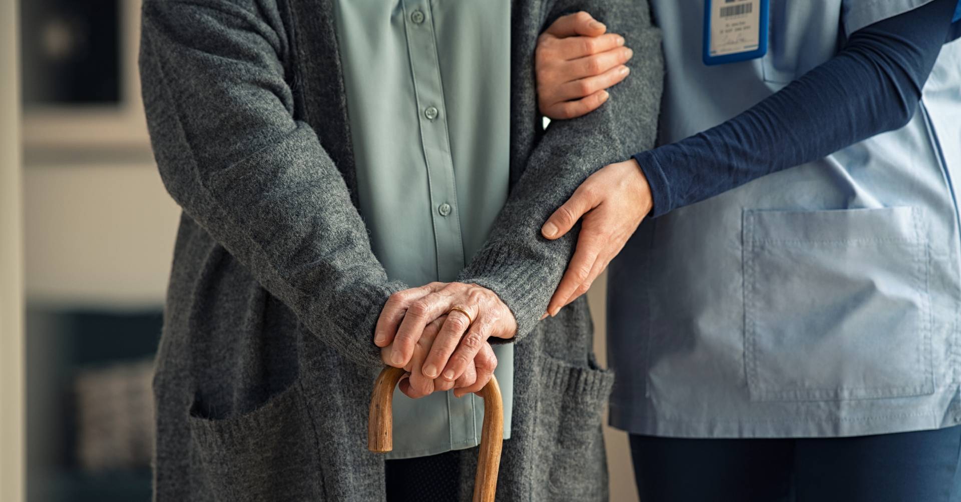 An elderly person, dressed in a grey cardigan, leans on a wooden cane for support. They are being assisted by a caregiver in a blue uniform, who gently holds their arm to offer stability and companionship.