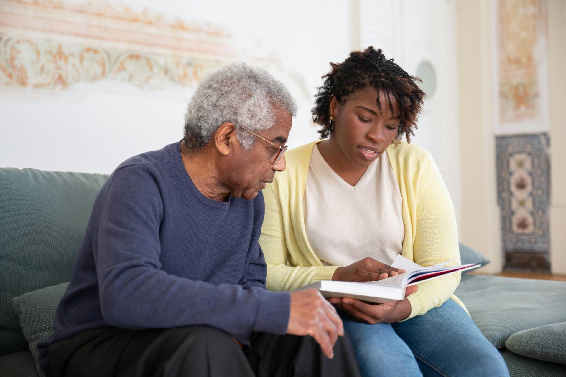 An older man with gray hair and glasses sits on a couch, reading a book with a younger woman beside him.