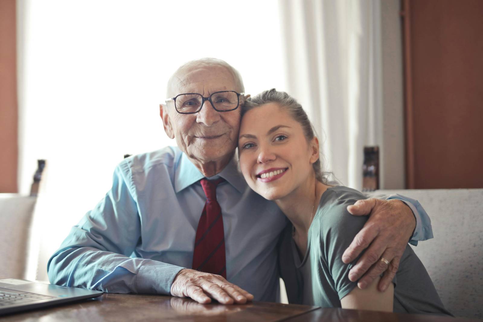 An elderly man in a light blue shirt and red tie embraces a young woman in a grey t-shirt. They are both smiling warmly at the camera while sitting at a wooden table in a well-lit room.