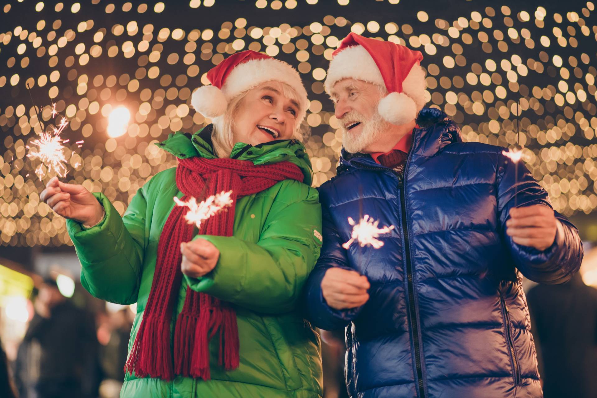 An elderly man and woman holding sparklers while standing in front of Christmas lights. They are wearing winter coats and Santa hats.