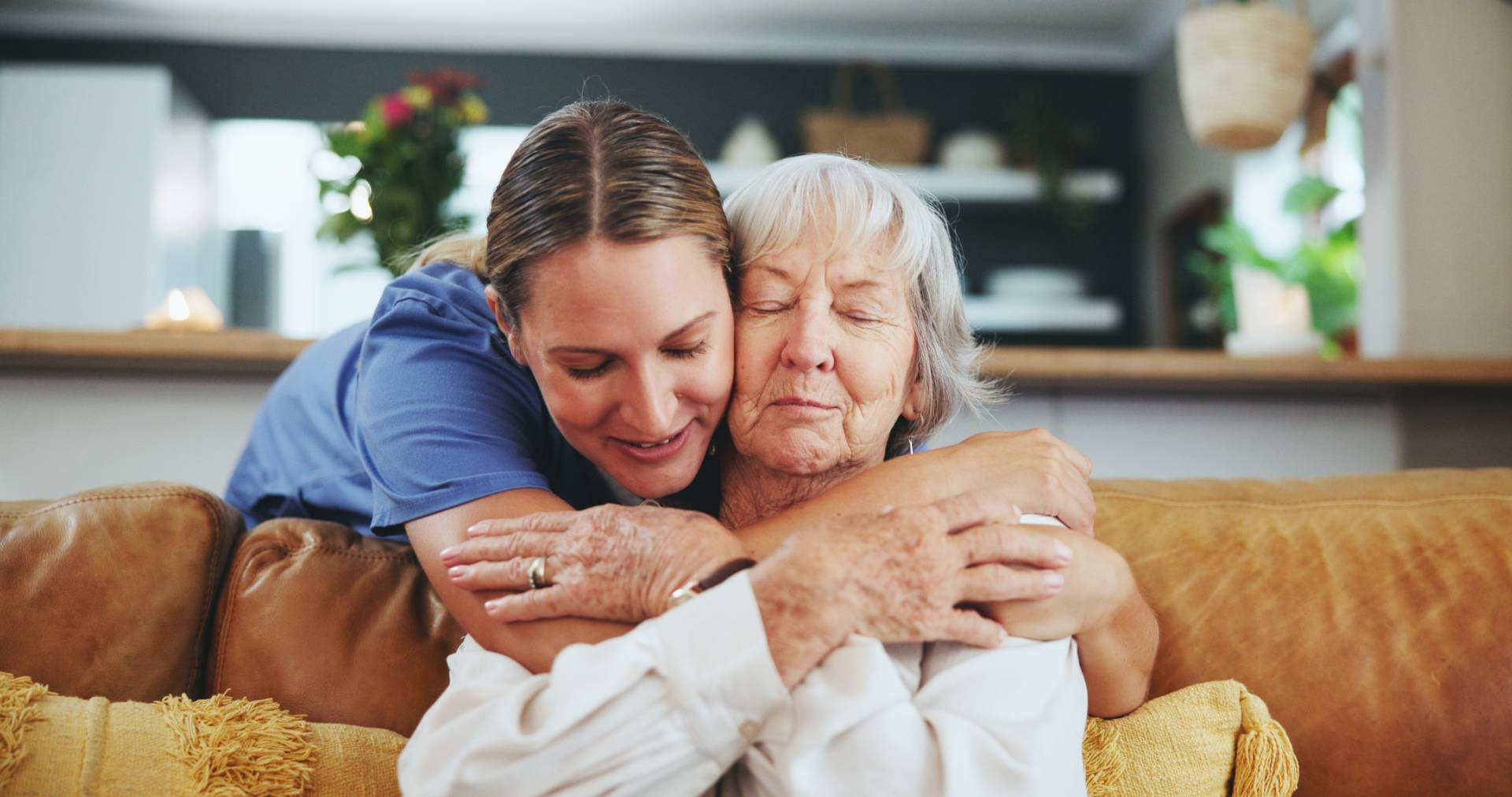 A caregiver hugging an elderly woman on a couch.