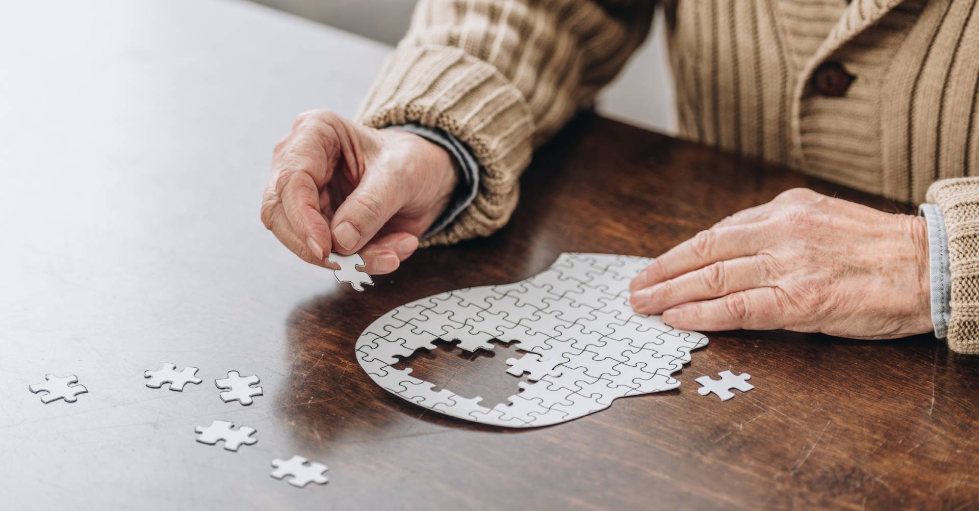 Close-up of an elderly person working on a puzzle shaped like a human head. The puzzle, made up of white pieces, symbolizes cognitive challenges, with several pieces missing around the brain area.