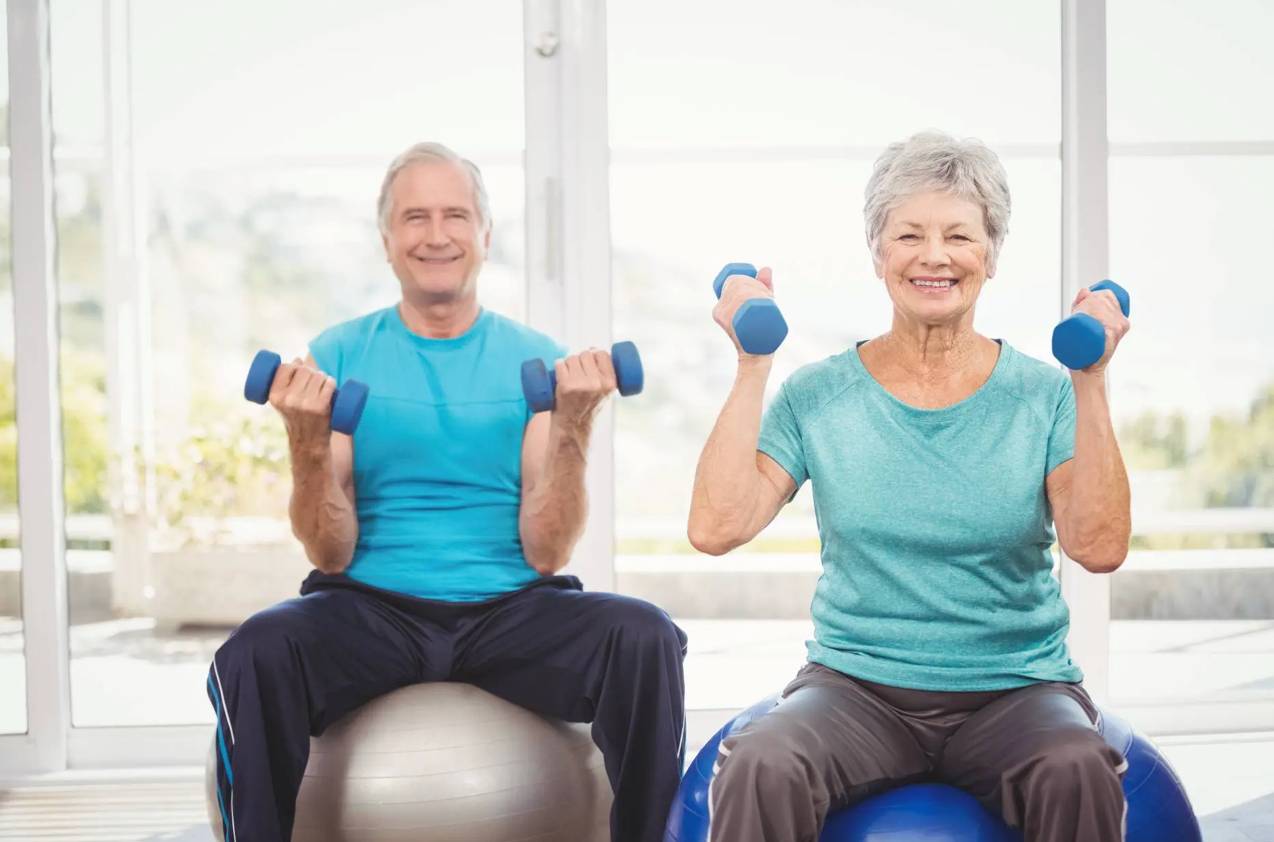 Elderly husband and wife sitting on a yoga ball lifting weights.
