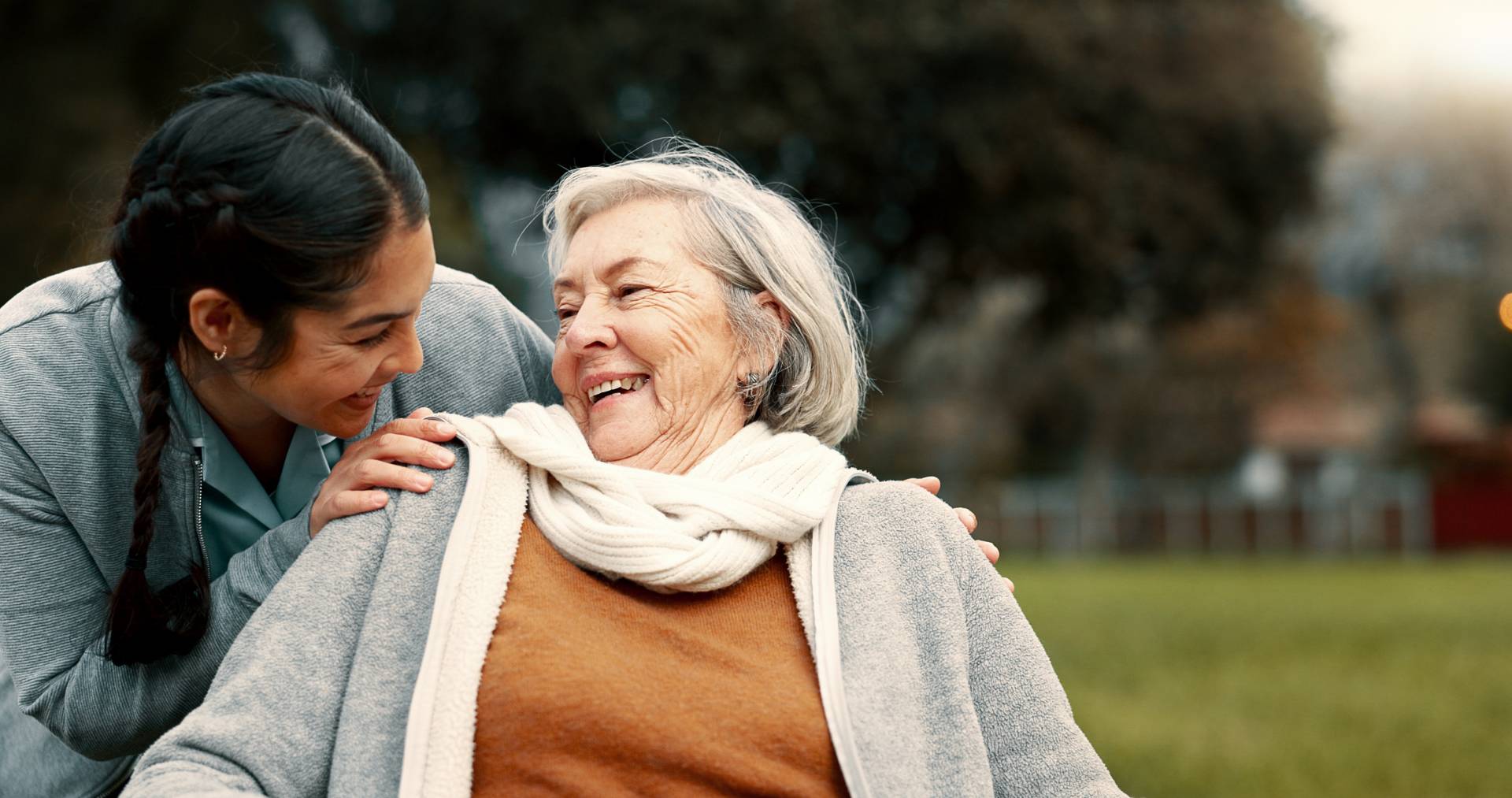 A joyful elderly woman, bundled in a scarf and cardigan, smiling and laughing with a caregiver.